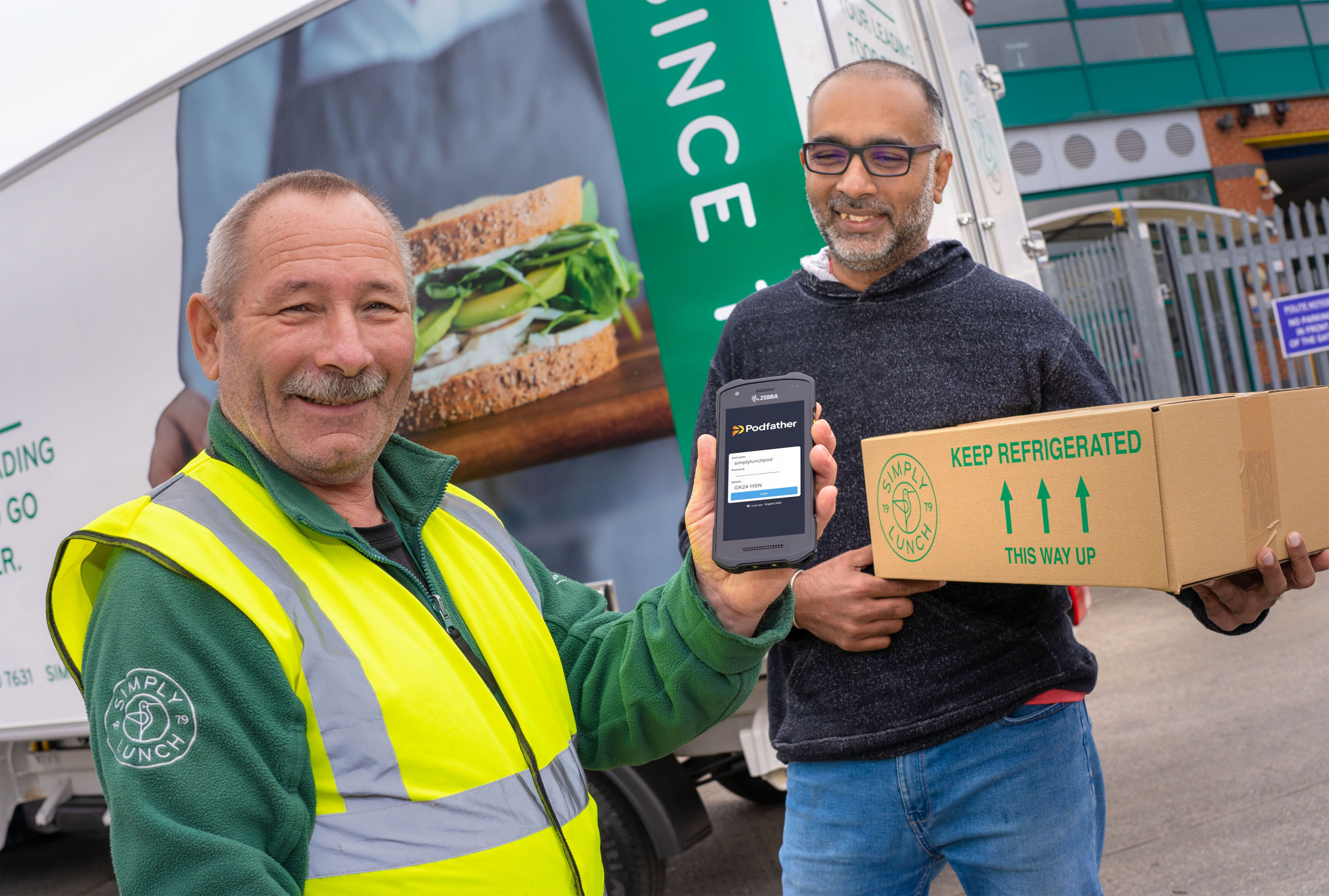 two men standing by a van; one is wearing a high vis vest and holding a mobile phone; the other is wearing a dark jumper and holding a box.