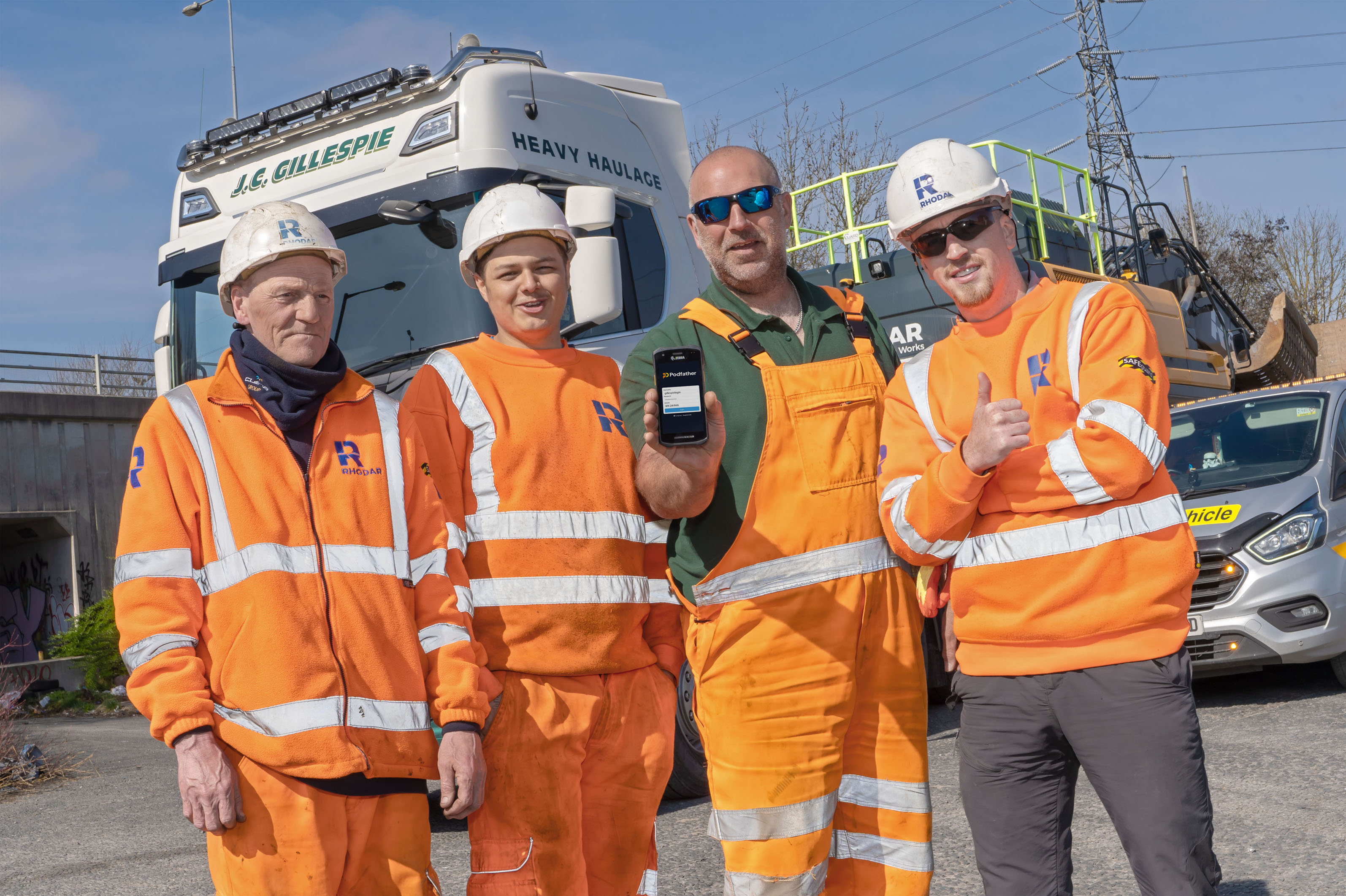 Four men standing in front of a lorry, with one holding a smartphone