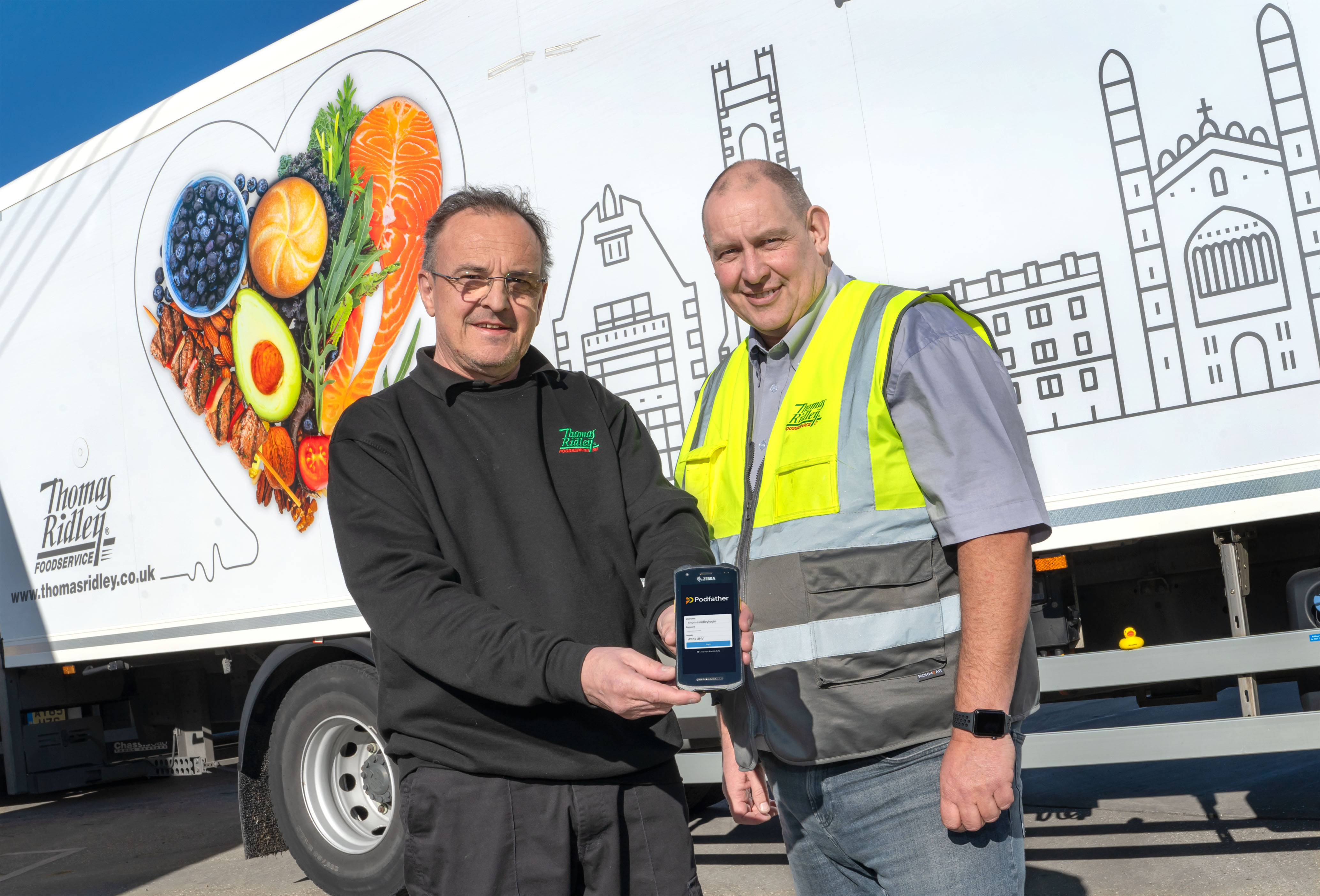Two men standing in front of a lorry, one holding a smartphone