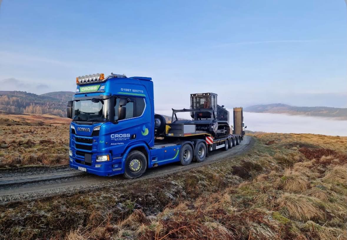 A blue heavy haulage truck carrying a digger in a countryside setting