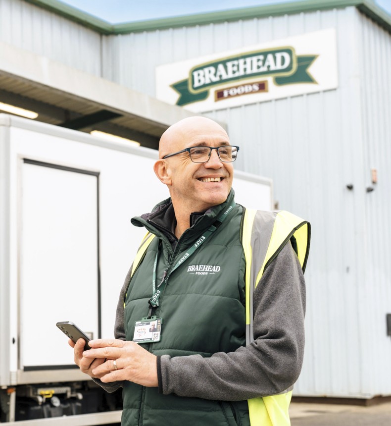 smiling man standing in front of a Braehead Foods depot holding a mobile device