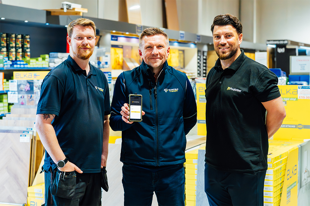 Three men in a warehouse smiling at the camera, one is holding a mobile device