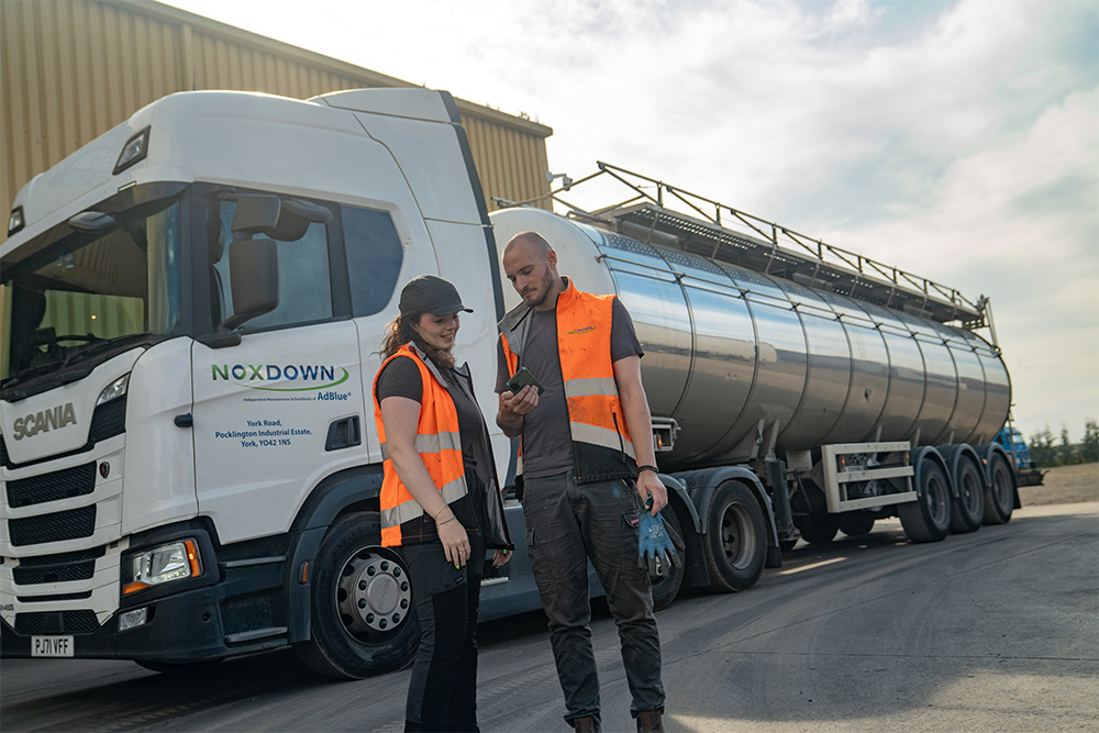 Man and woman standing in front of a silver tanker vehicle looking at a mobile device