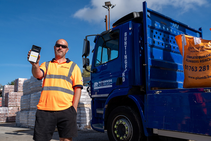 Man holding a mobile device wearing an orange high vis shirt standing in front of a blue truck.