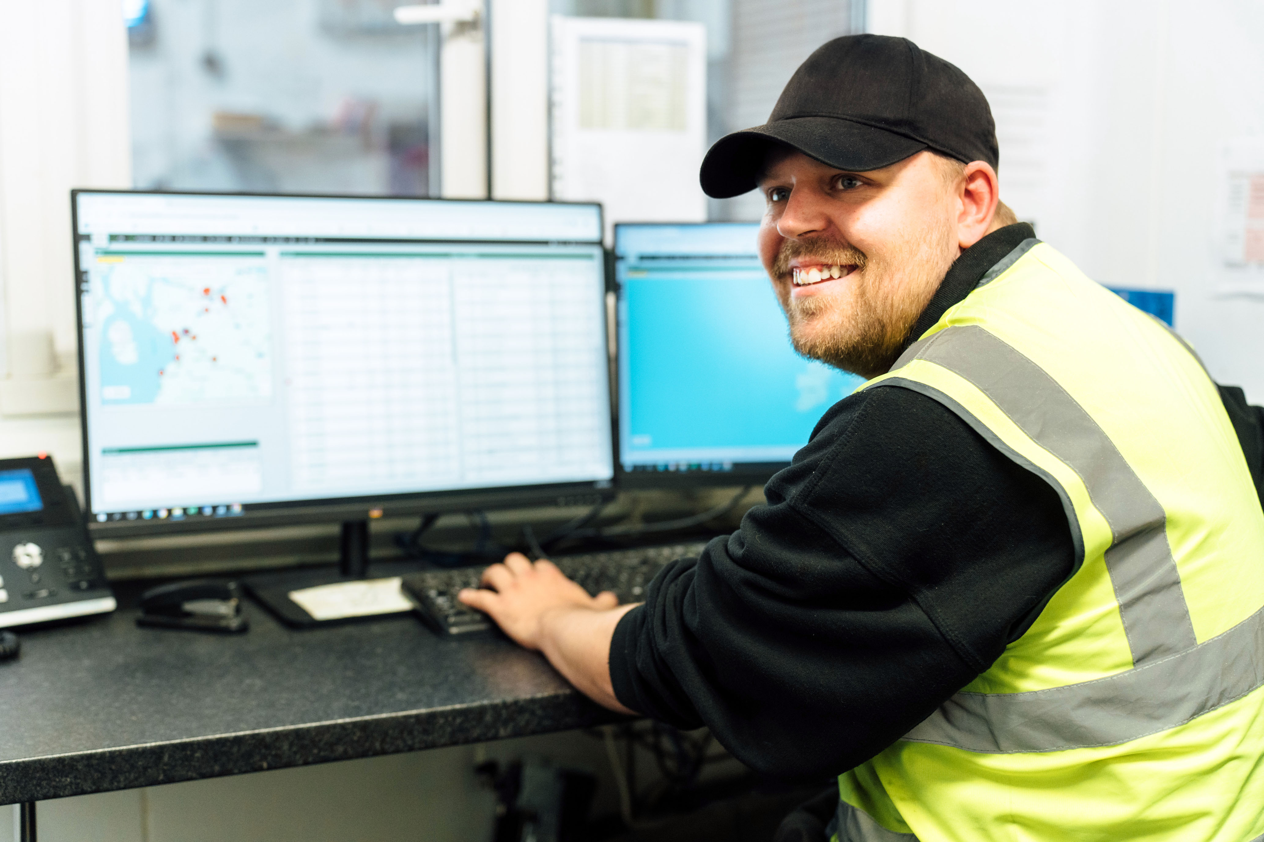 smiling man wearing a high vis vest and a cap sitting at a computer